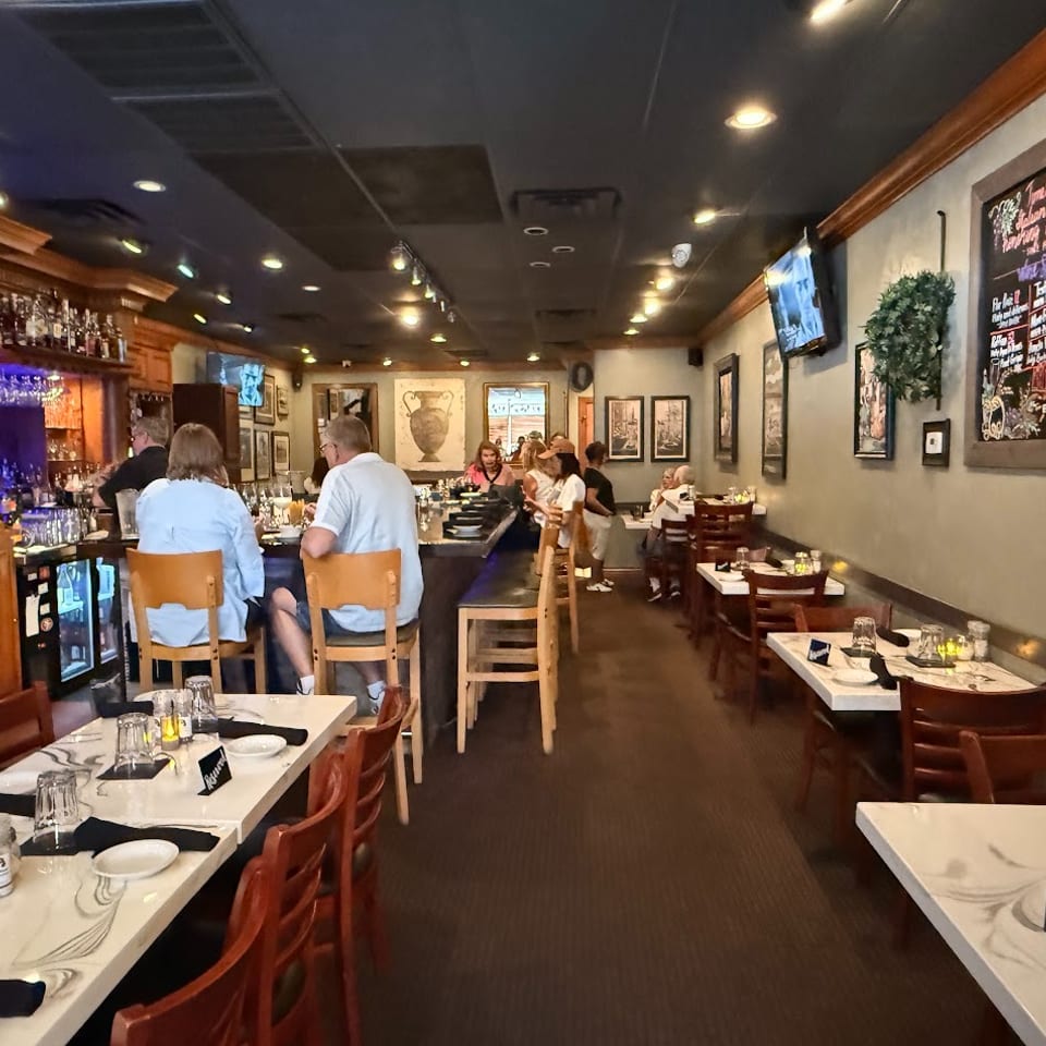 Warmly lit restaurant dining room with a full bar, seated diners, set tables, framed artwork, and a chalkboard menu.